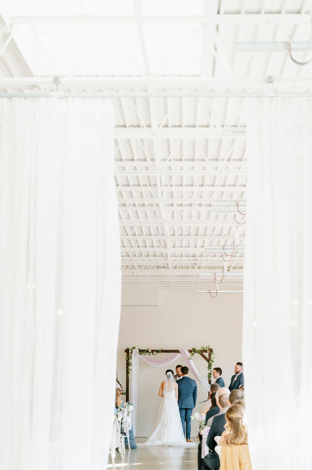 Wedding ceremony taking place in a bright white industrial venue with exposed ceiling beams and flowing curtains.