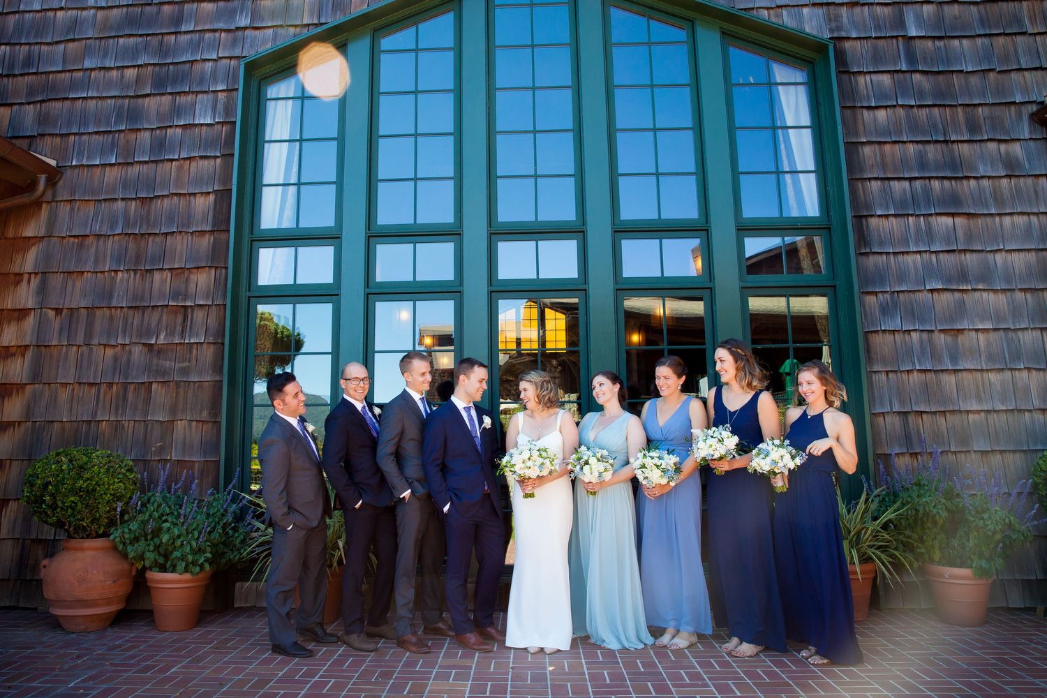 Wedding party in dark suits and formal attire laugh together while holding white floral bouquets outdoors.