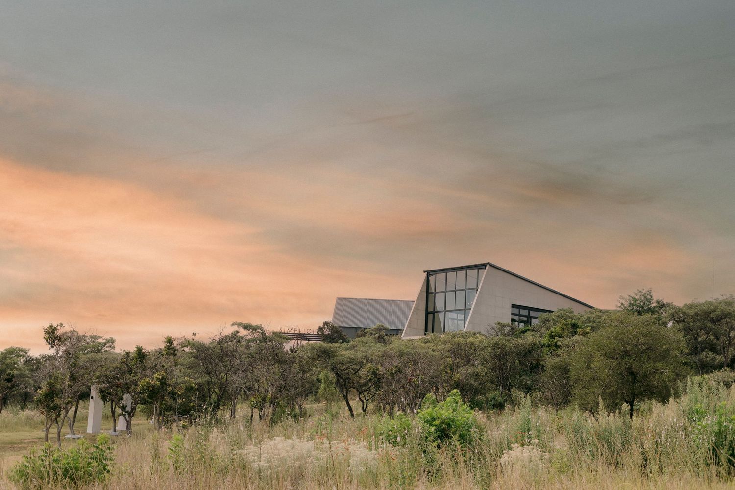 Modern angular building surrounded by African bushveld under a dramatic orange sunset sky.