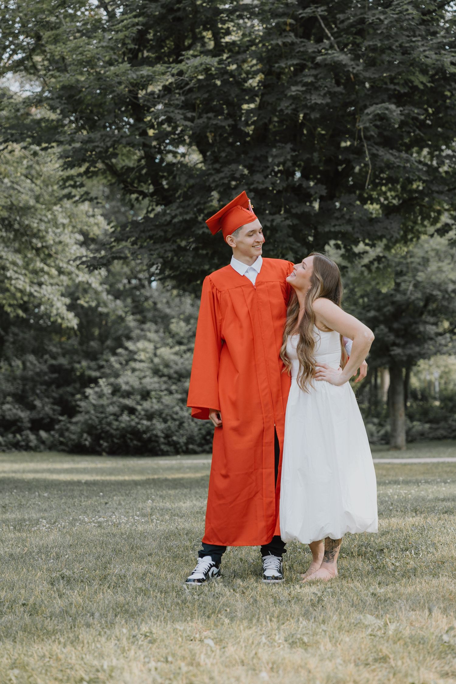 Graduate in red cap and gown smiling with a girl in a white dress in a green park setting.