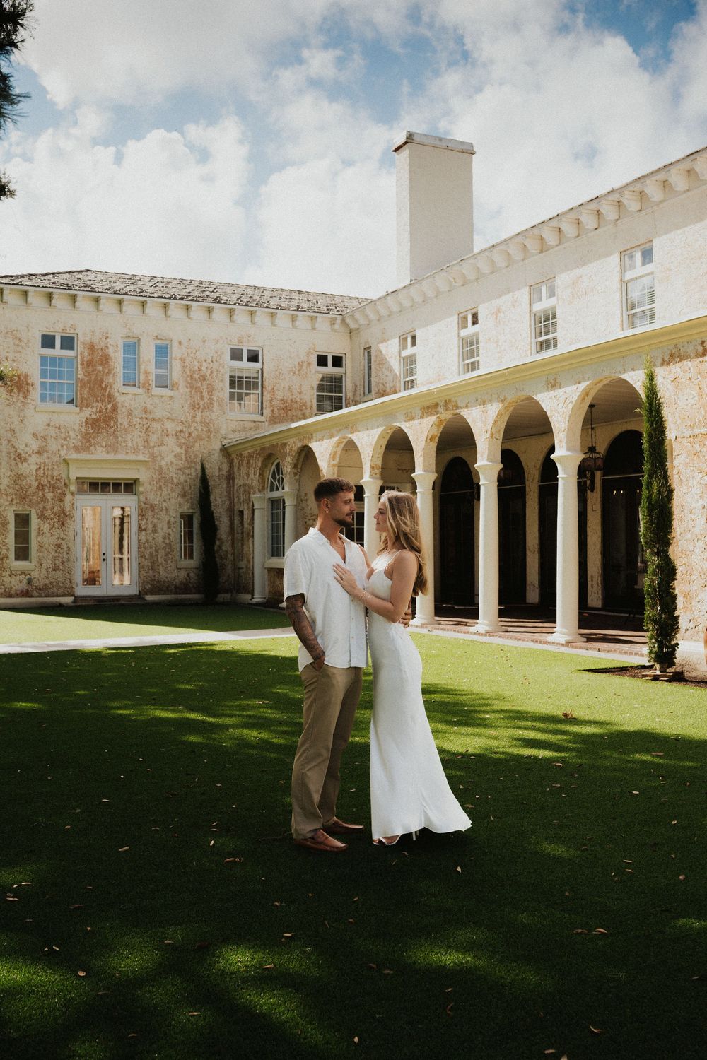 A couple dances on a manicured lawn surrounded by elegant stone archways and columns of a historic building.