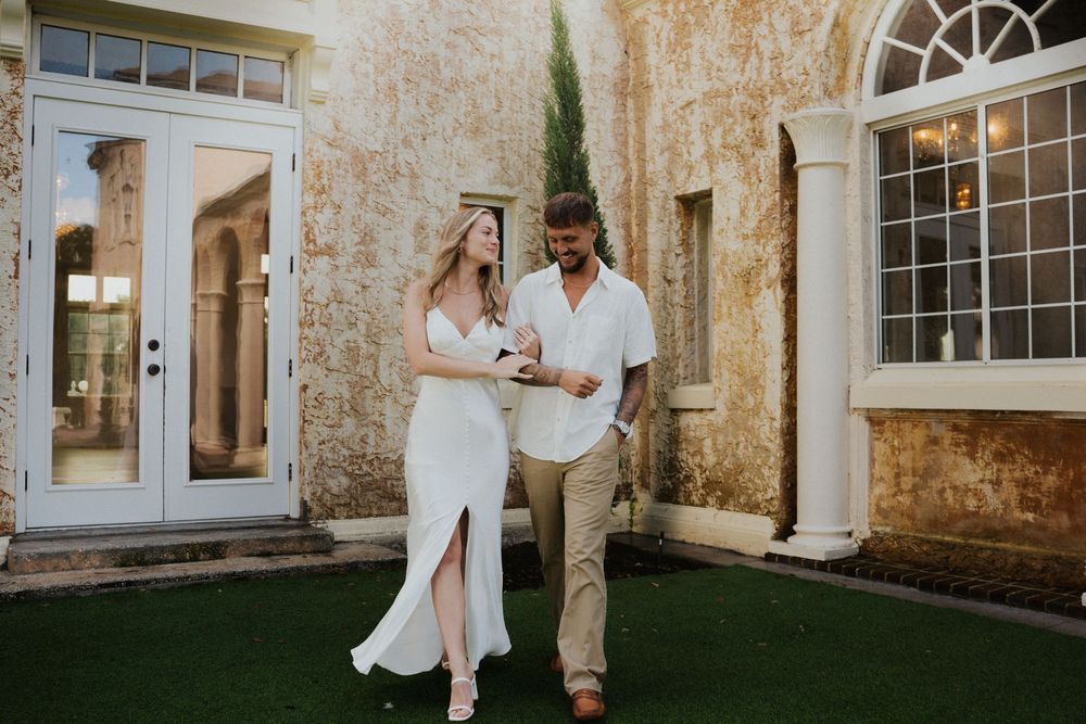 A couple shares a tender moment on manicured grass in front of a historic stone building.