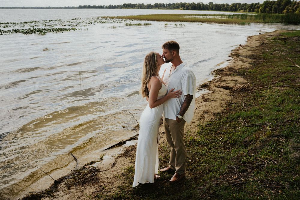 A couple embraces on a rocky shoreline with water and natural landscape in the background.