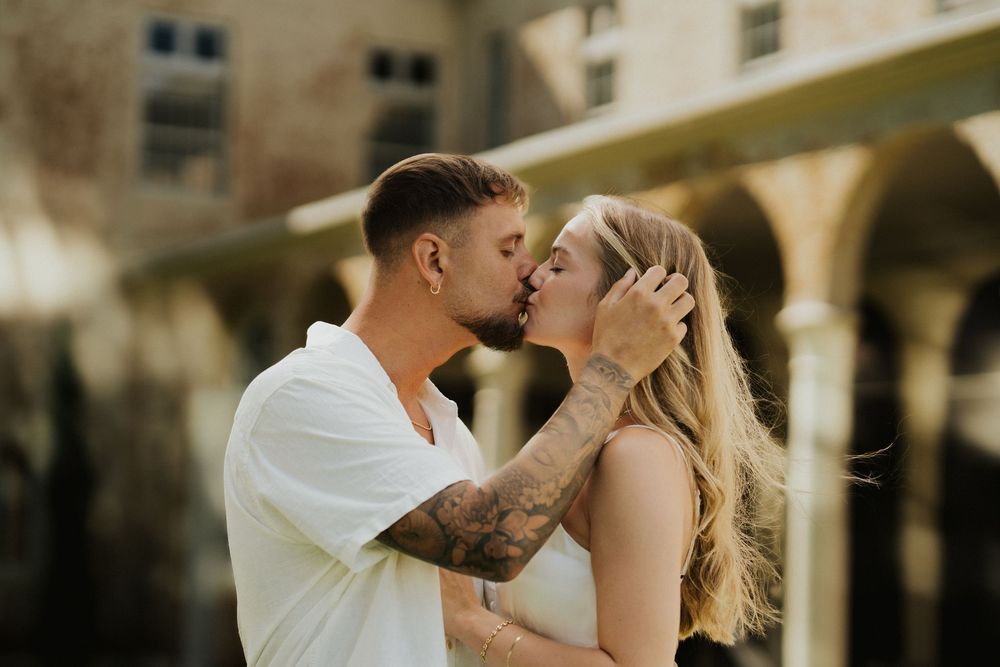 A romantic couple shares a passionate kiss while standing outdoors in a sunlit setting.