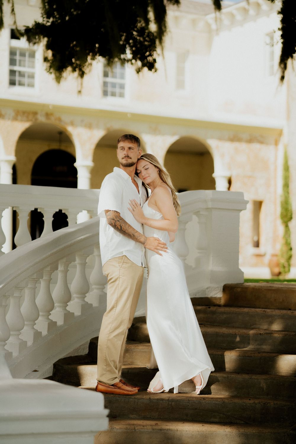 A romantic couple in wedding attire poses on white stone steps with elegant columns and arches in the background.