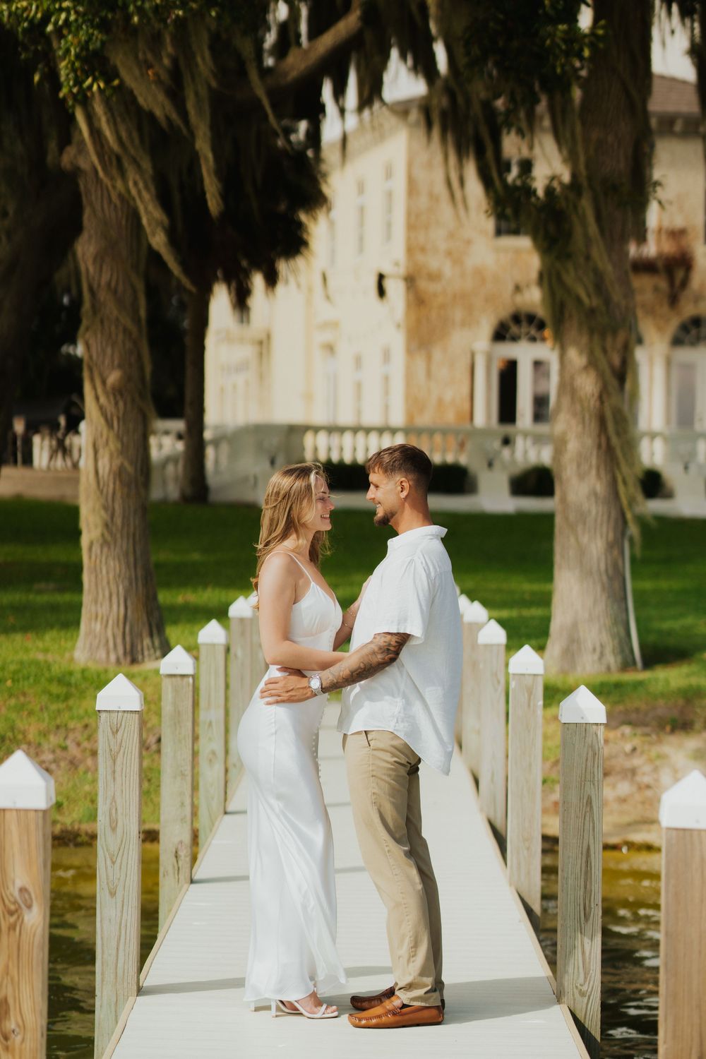 A romantic couple stands together on a white wooden dock surrounded by trees and a historic building.