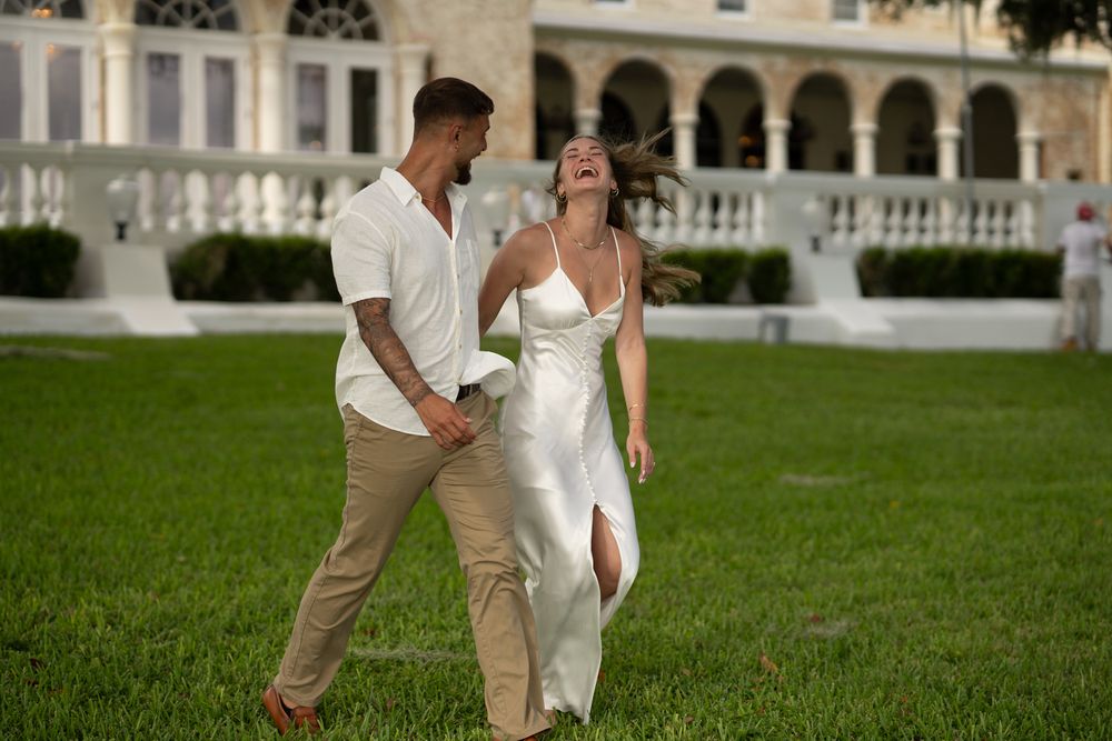 A couple in white and beige outfits walk hand in hand on a grassy lawn near a grand building.