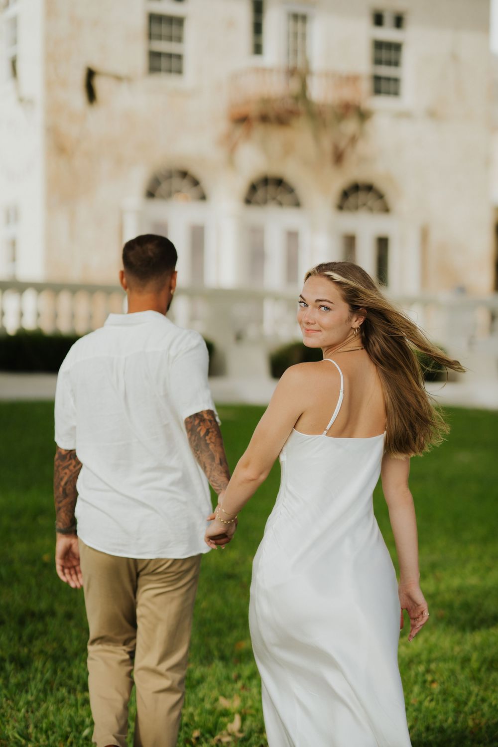 A couple walking hand in hand on grass in front of a grand white building with arches.