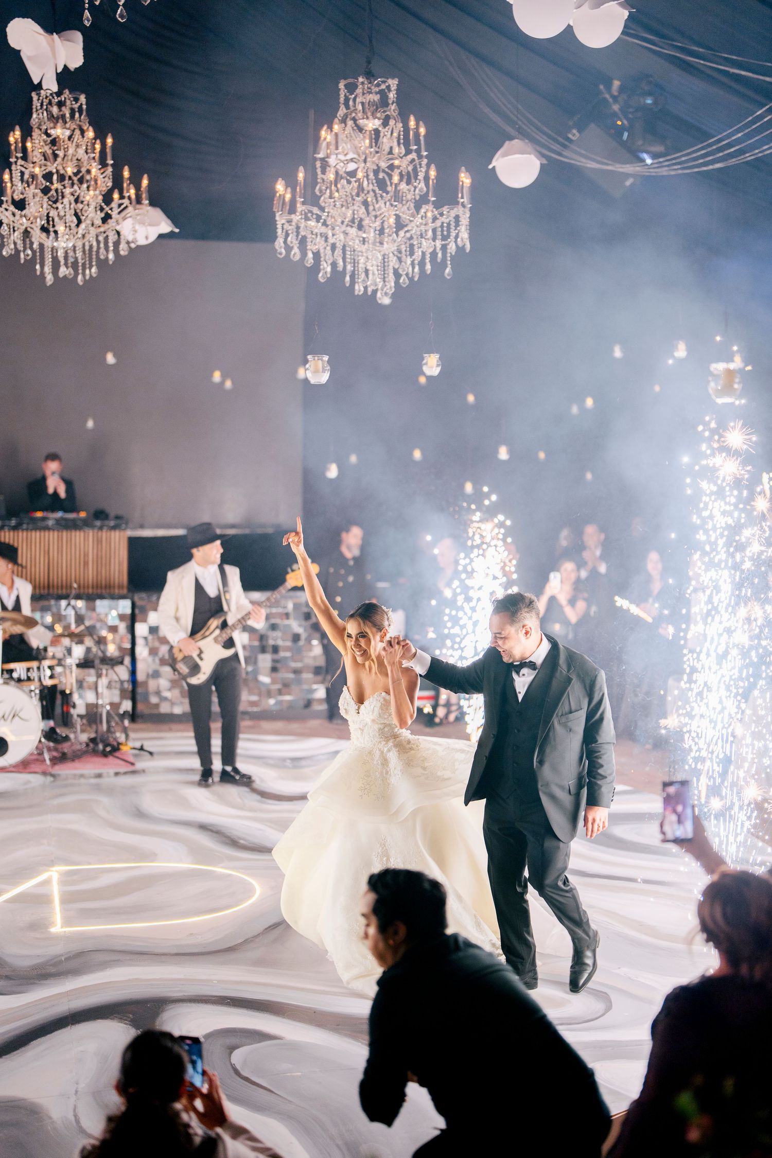 Elegant wedding reception with crystal chandeliers and sparklers on white dance floor during couple's first dance.