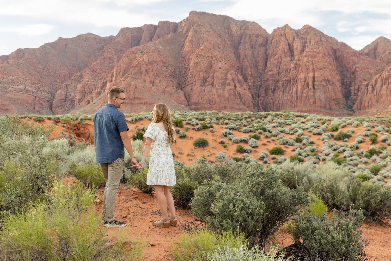 Couple holding hands in red rock desert landscape with sage brush and dramatic sandstone cliffs in background. Ivins utah