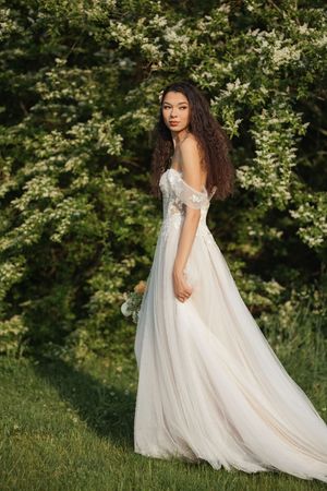 Ethereal bride in flowing white gown poses gracefully in a lush garden setting during a summer photoshoot.