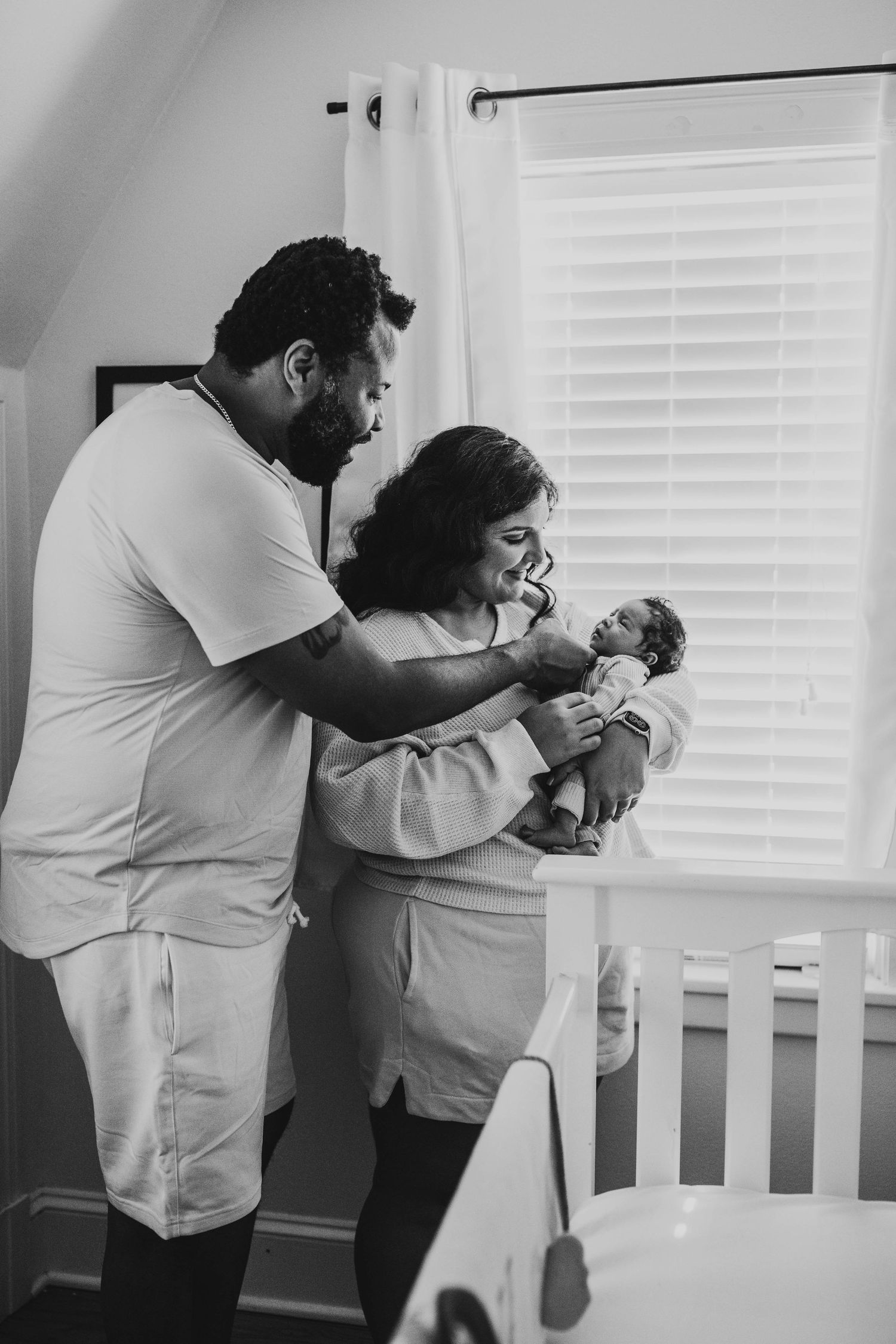 A couple in white clothing admires their newborn baby near a crib in a black and white photo.