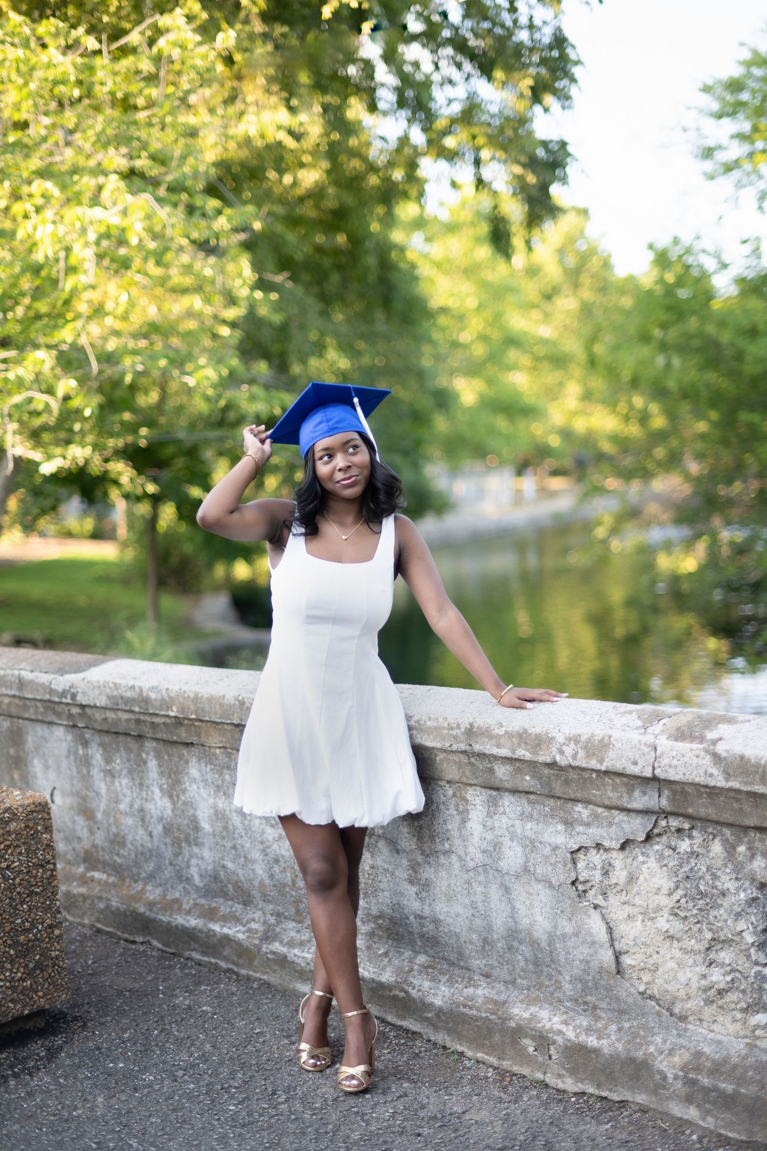 A graduate in a white dress and blue cap poses playfully on a stone bridge in a sunny park setting.