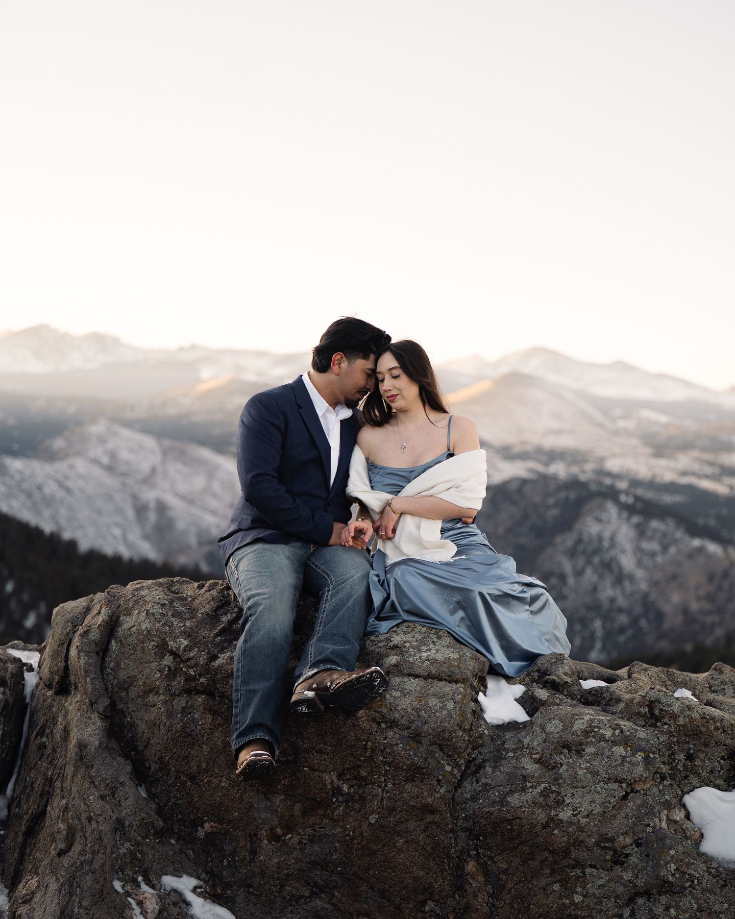 A couple shares an intimate moment while sitting on rocky cliffs overlooking a mountainous landscape at sunset.
