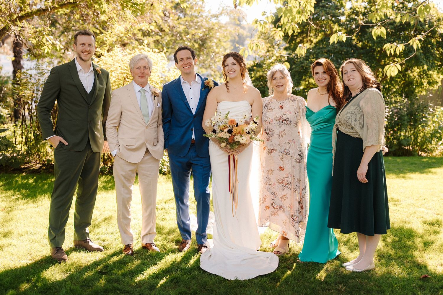 Wedding party poses in a row outdoors with sunlit trees and greenery in the background.