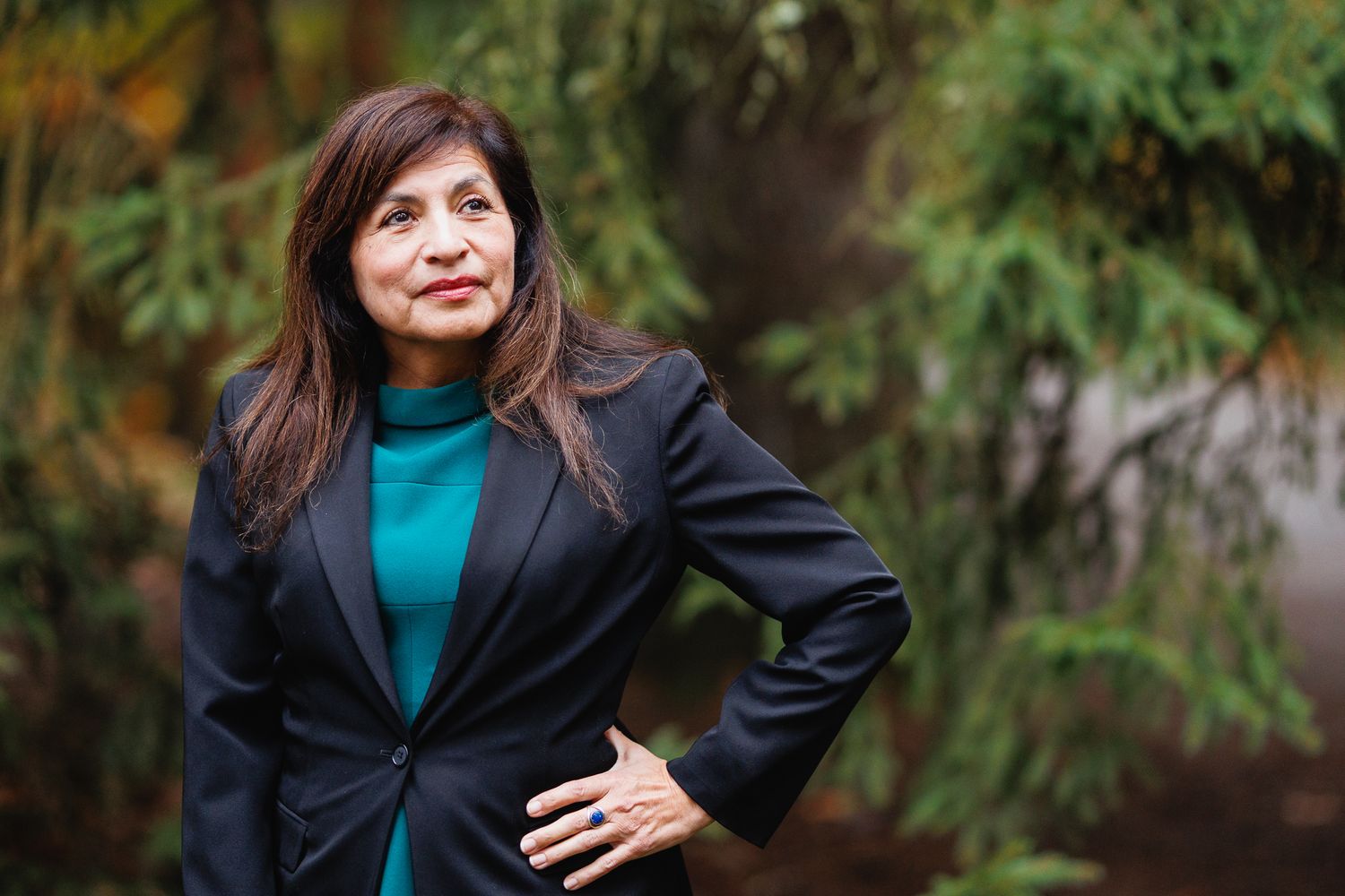 Professional woman in teal blouse and navy blazer poses confidently outdoors with green foliage background.