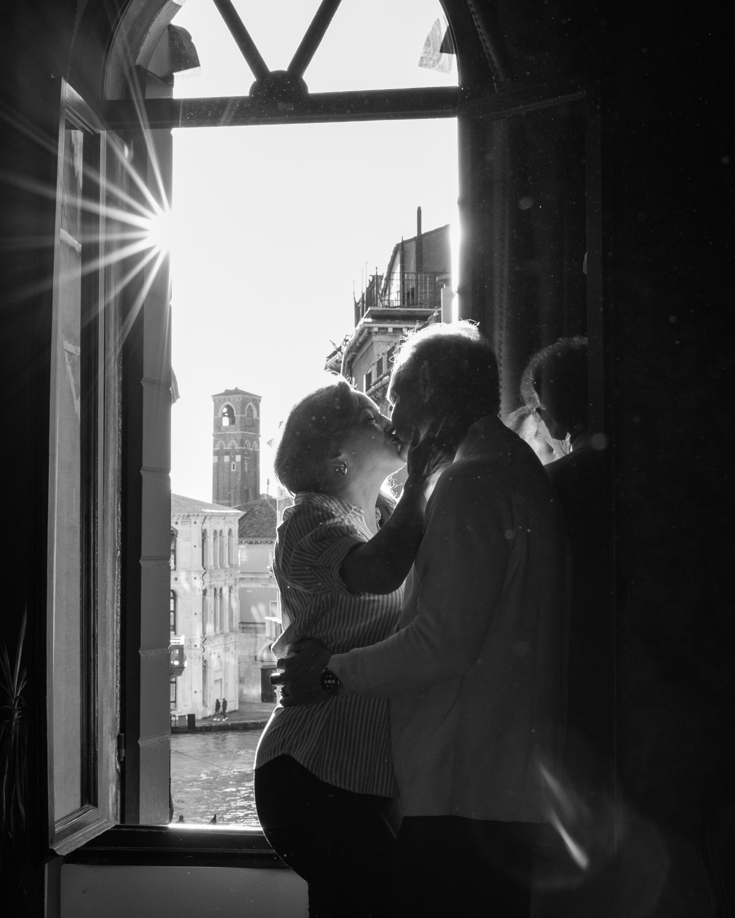 A silhouetted couple gazes out of an open window at historic Italian architecture with sunlight streaming through.