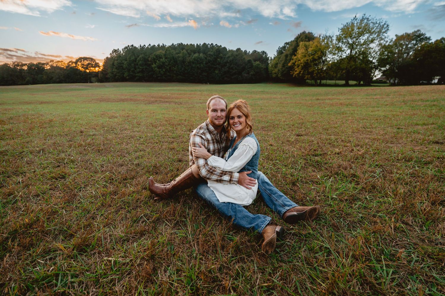 A couple sits together in a grassy field during golden hour as the sun sets behind trees in the distance.