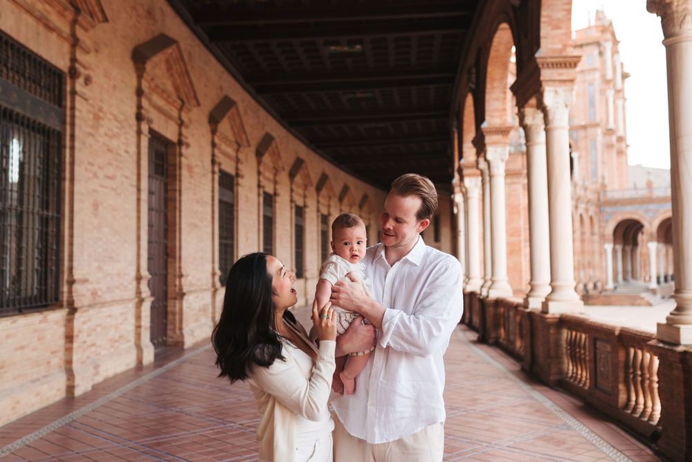 A couple shares a joyful moment holding their baby in a stunning Spanish arcade with ornate arches.