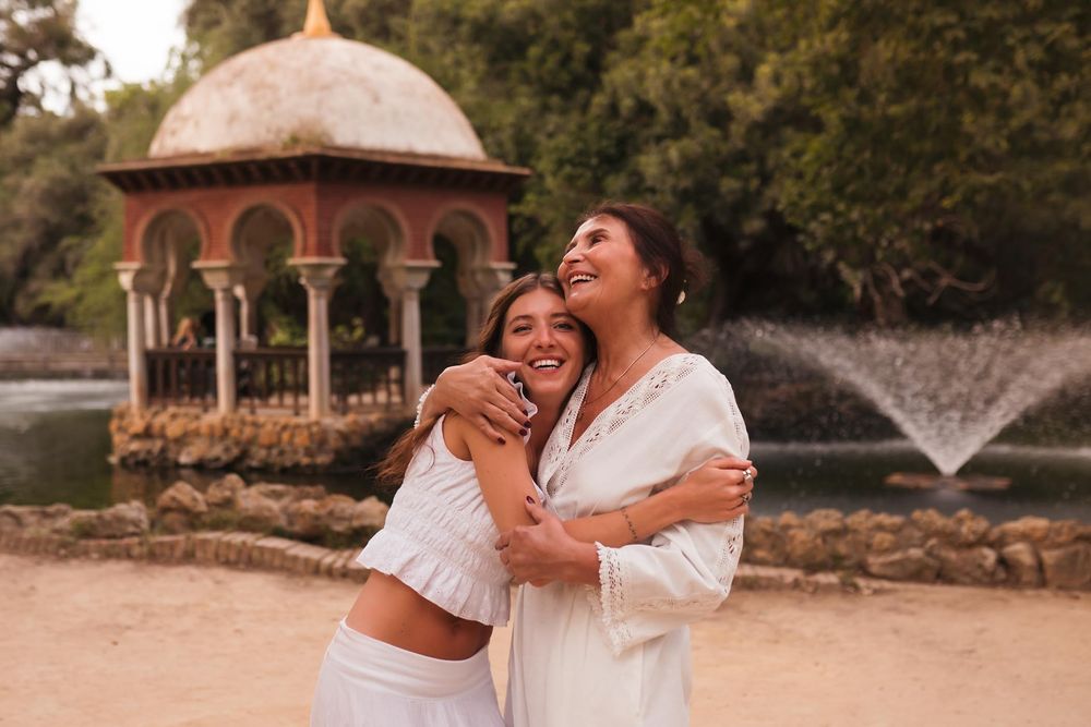 Two women in white outfits hugging and laughing in a park with a fountain and pavilion backdrop.