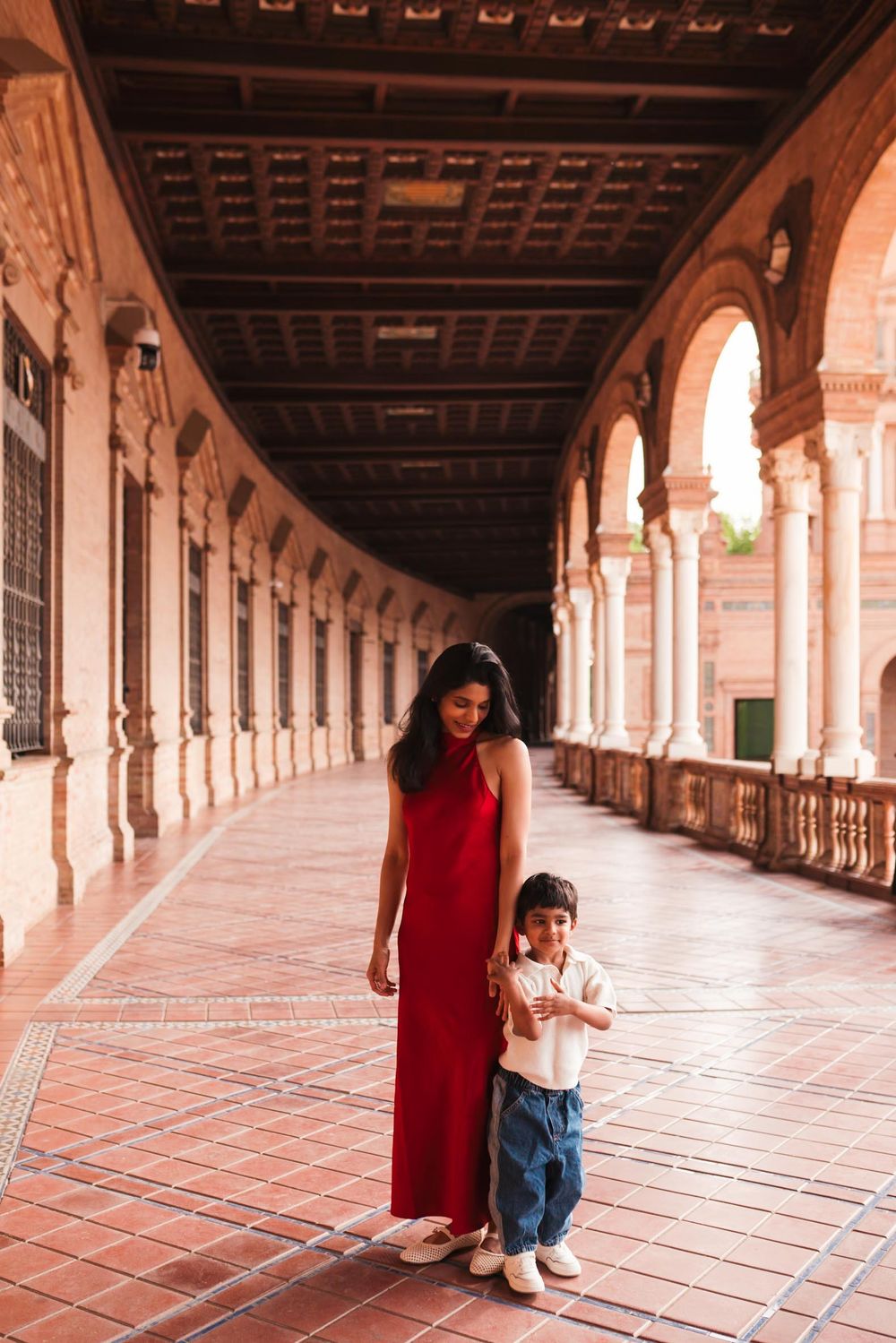 Woman in red dress and child standing in ornate Spanish arcade corridor with arches and terracotta tiles.