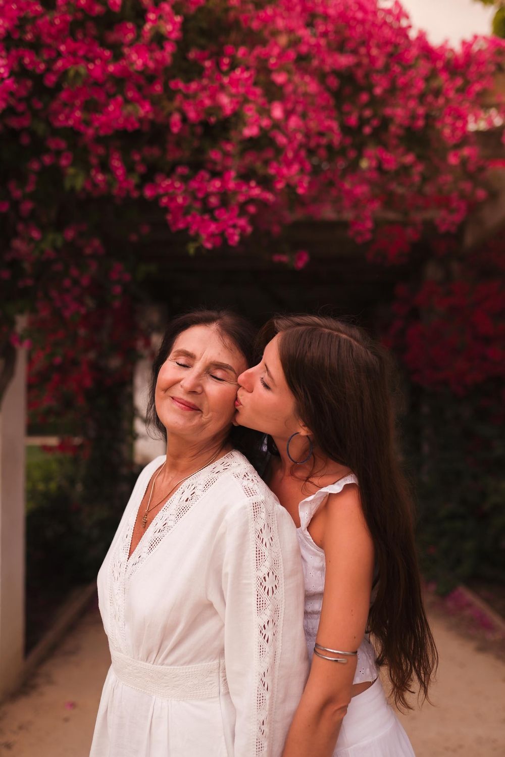 Young woman kissing older woman on cheek, both in white dresses, surrounded by vibrant pink bougainvillea flowers.