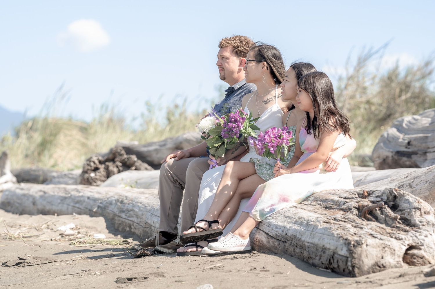 A couple relaxes on a sandy beach near driftwood logs on a sunny afternoon.