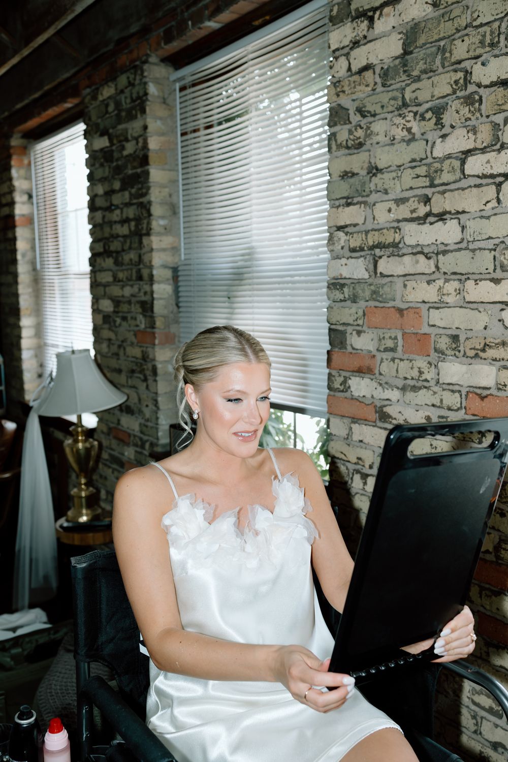 A person in white dress reads from a folder while seated against exposed brick wall.