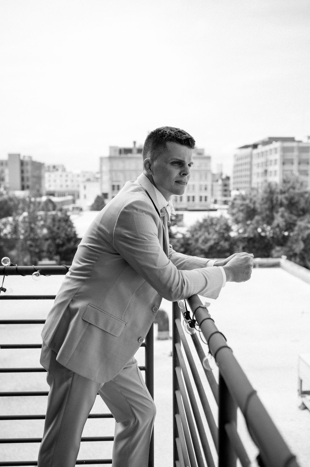 Black and white photo of a person in formal attire leaning on an outdoor railing with city buildings in background.