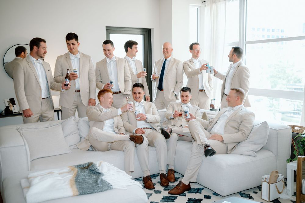 Groomsmen in light colored suits relax on white furniture during wedding preparations.