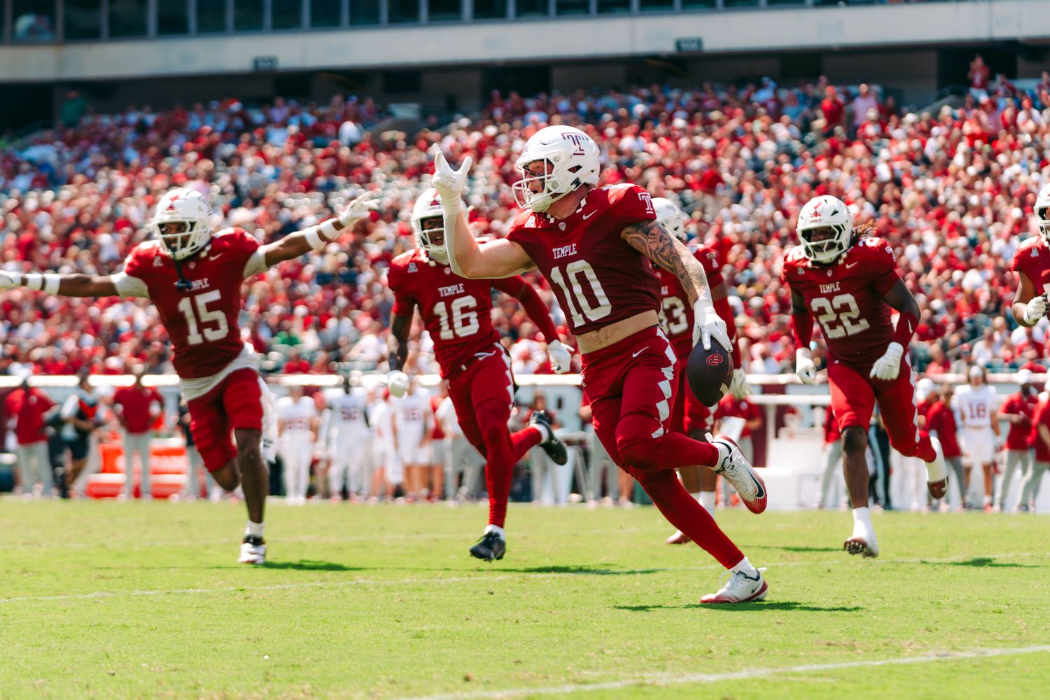 Football players in red uniforms execute a running play during a game with a packed stadium in the background.