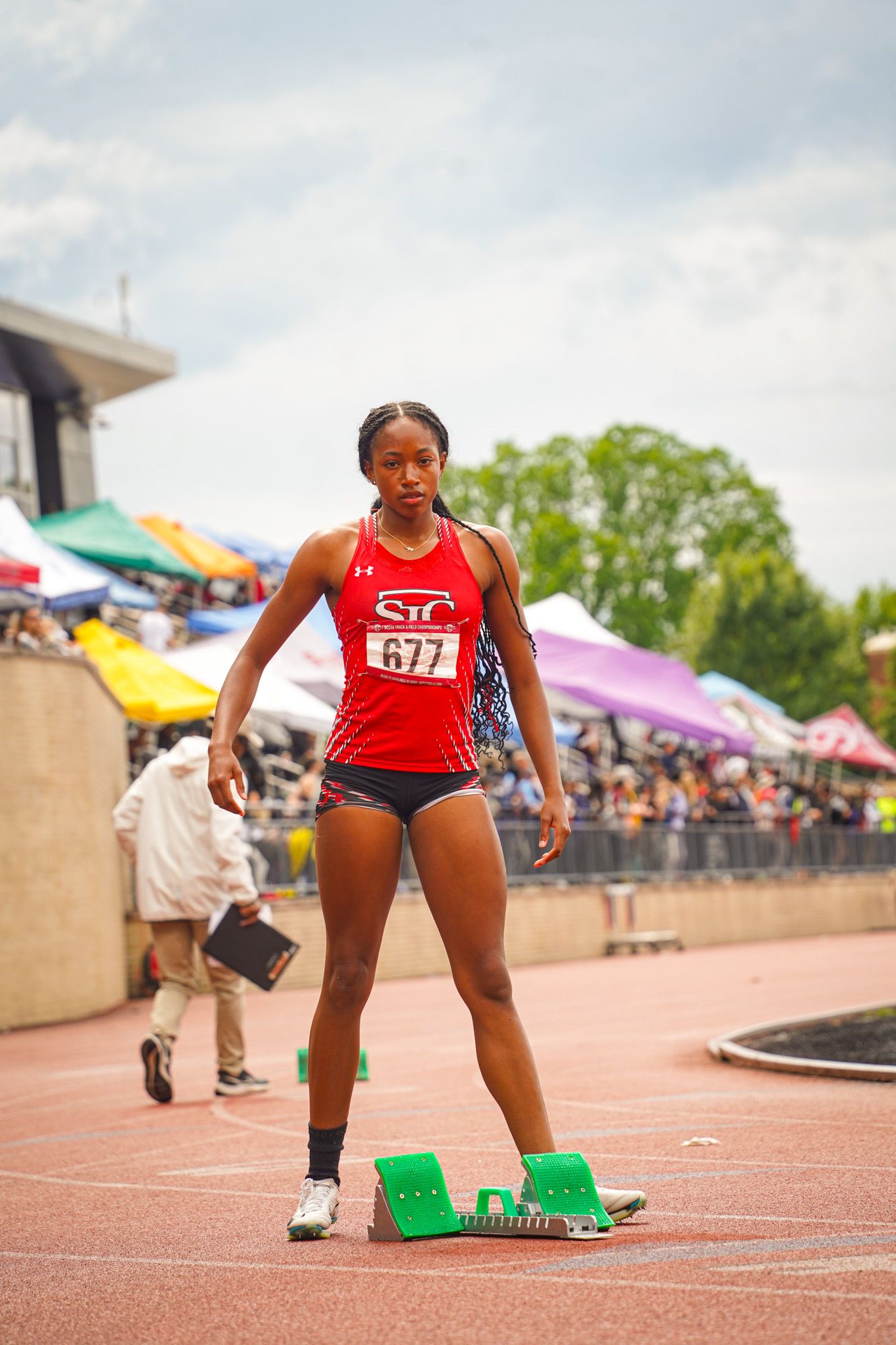 Track athlete in red uniform crouches at starting blocks preparing for race at outdoor stadium competition.