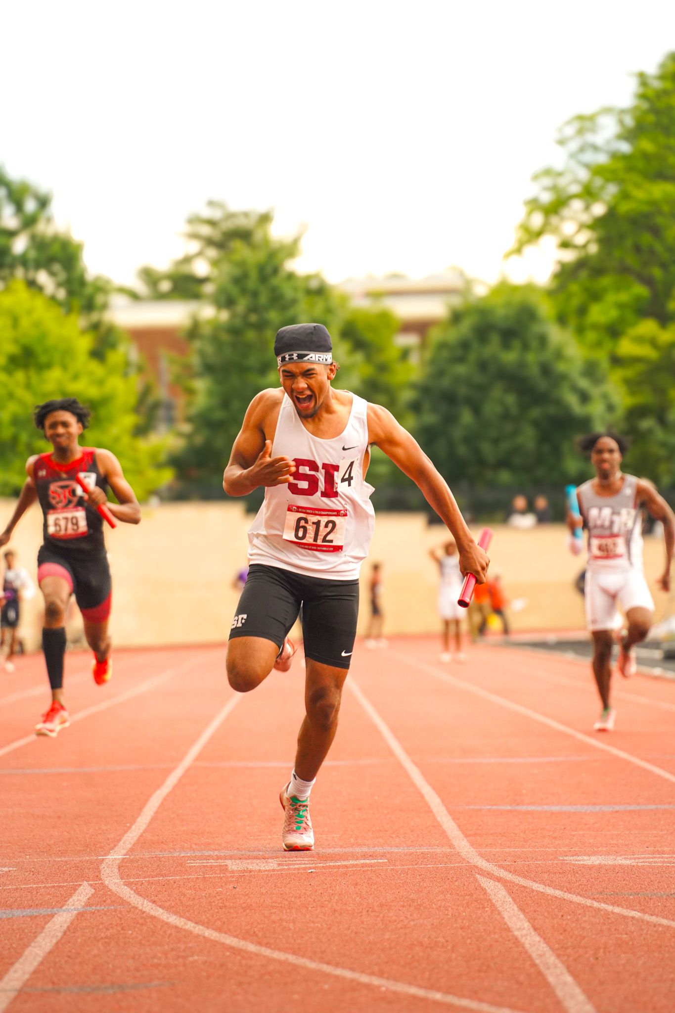 Track athletes compete in a sprint race on a red running track during an outdoor competition.