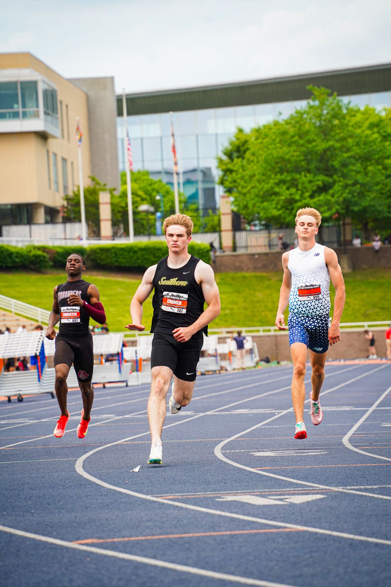 Track athletes compete in a sprint race on an outdoor running track with buildings and trees visible in the background.