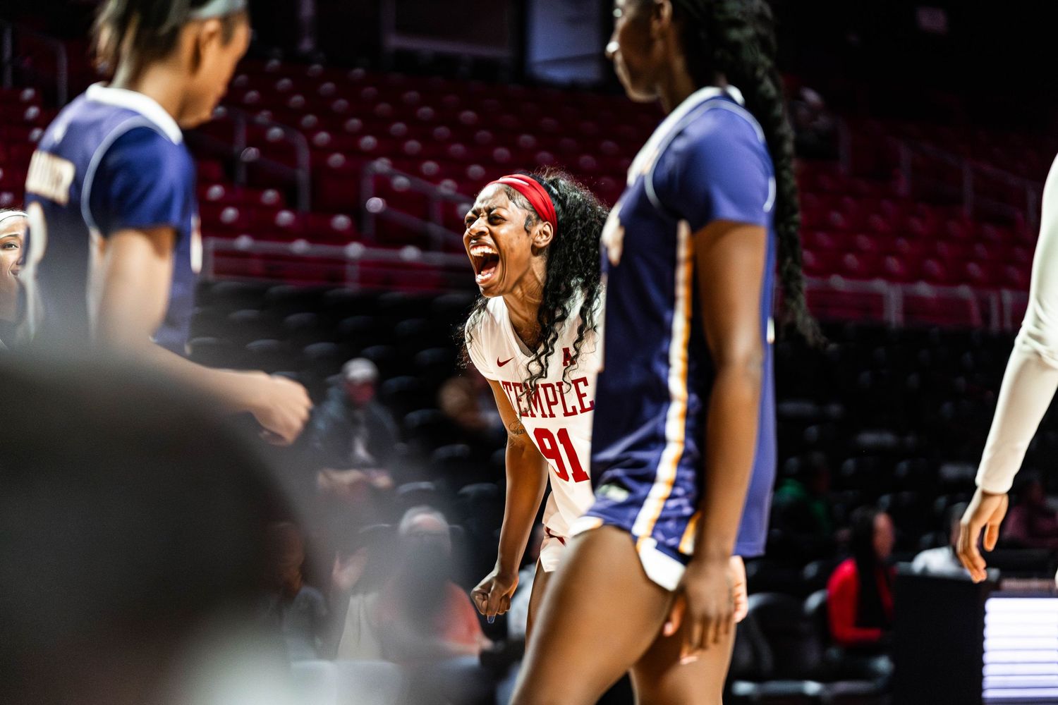 College basketball player celebrates during a game against opponents wearing blue uniforms in an arena with red seats.
