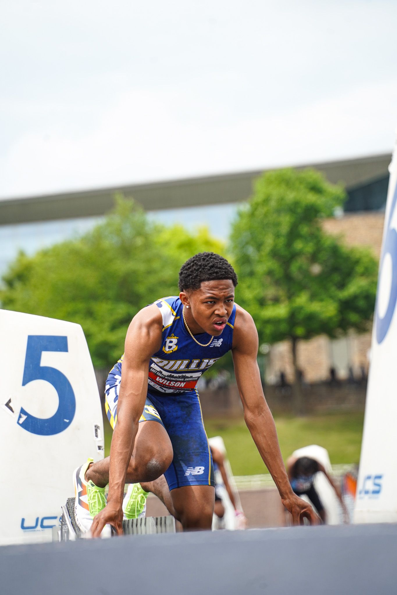 Track athlete in blue uniform crouches at starting position on lane 5 during outdoor competition.