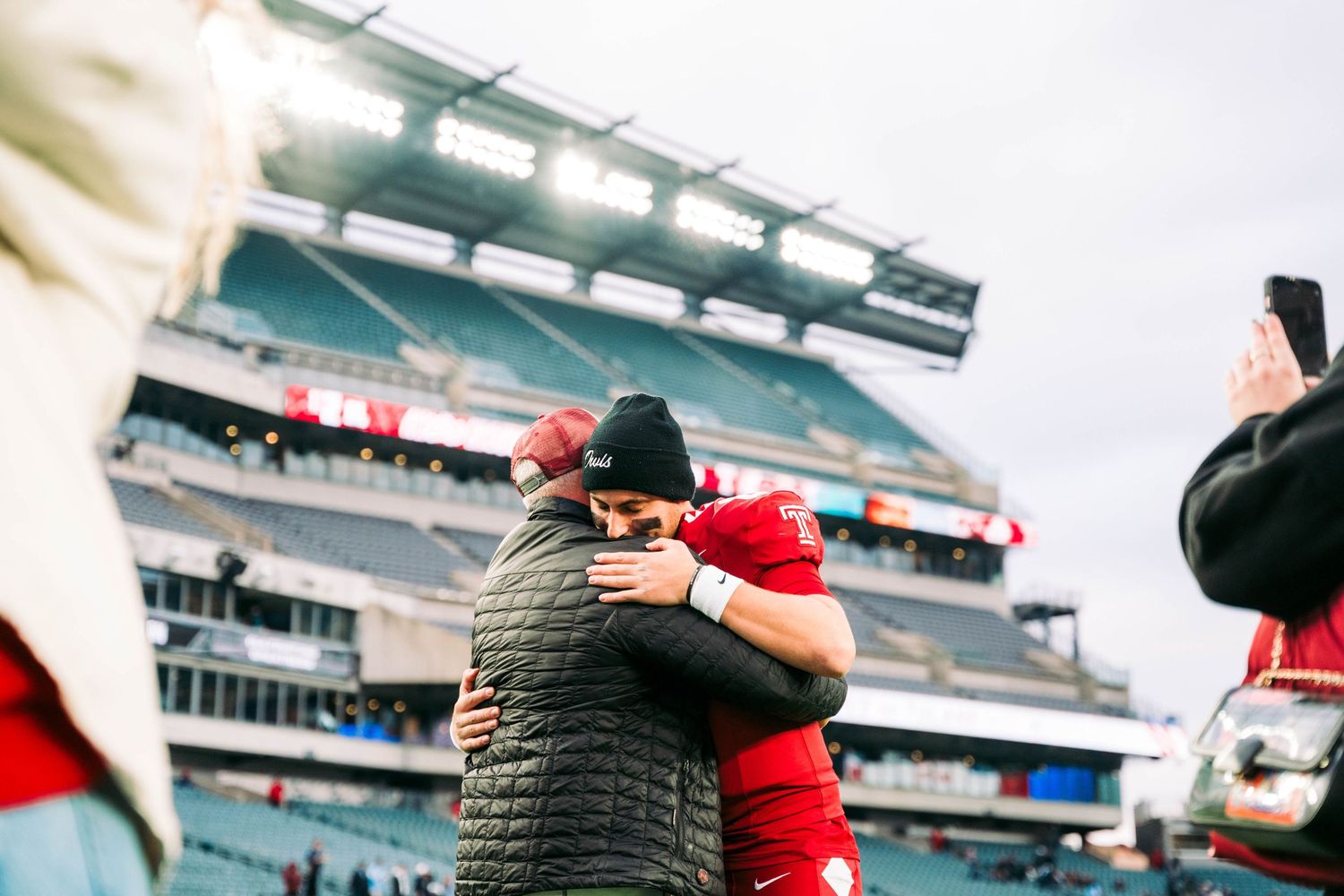 Two people share an embrace while wearing winter coats at a football stadium on an overcast day.