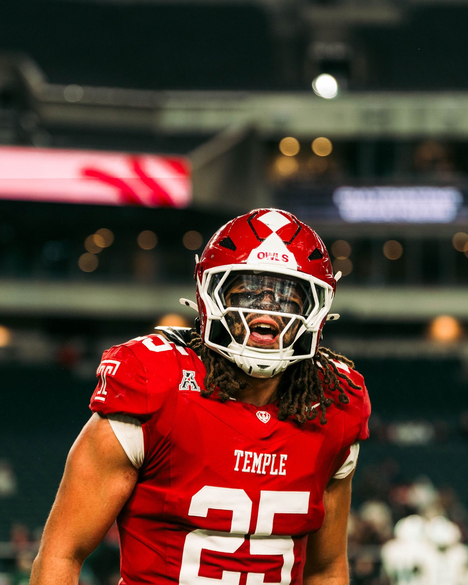 Temple football player in red uniform number 25 celebrates on field with stadium lights in background.