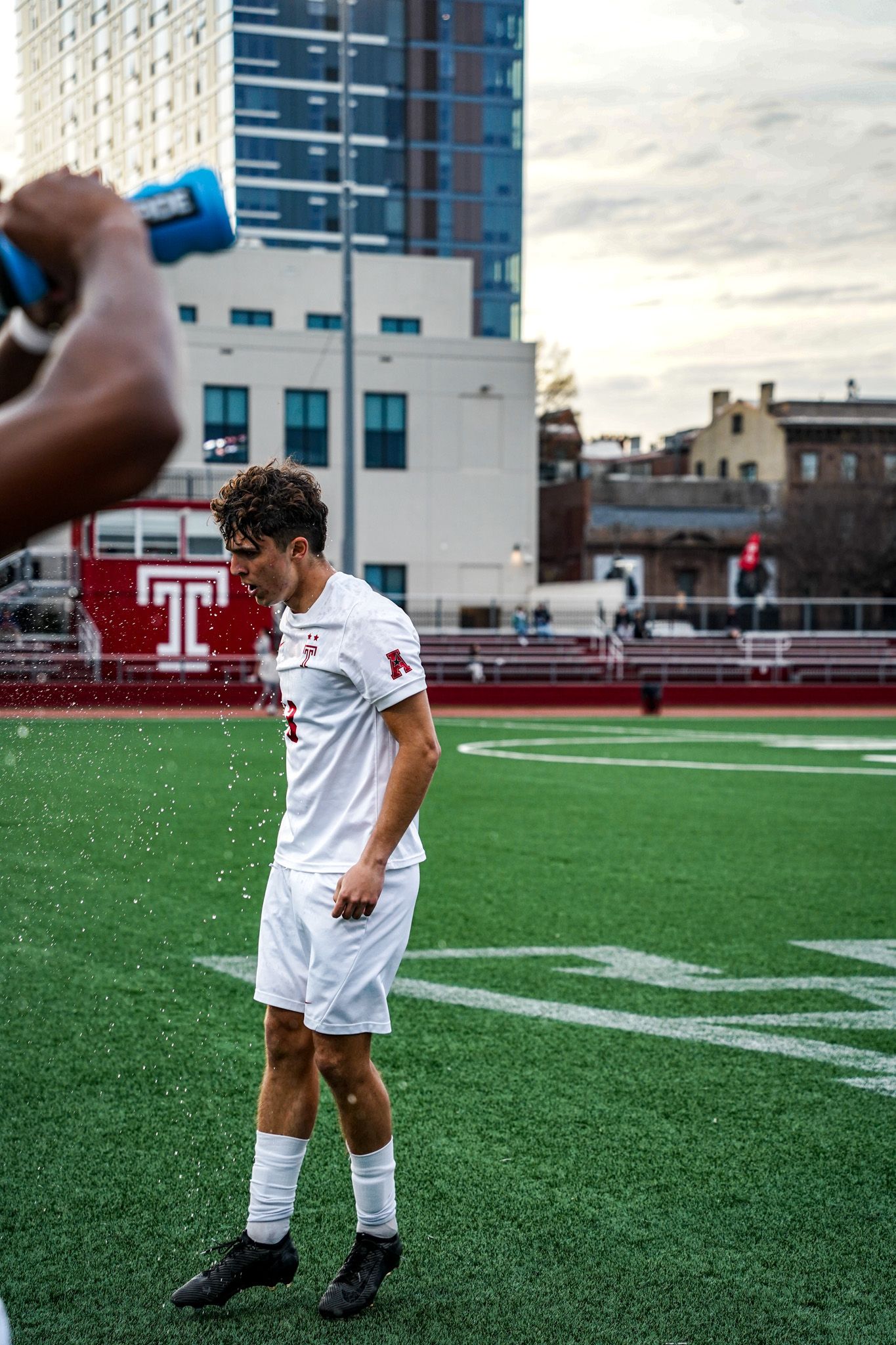 Soccer player in white uniform practices footwork drills on artificial turf field with stadium buildings in background.