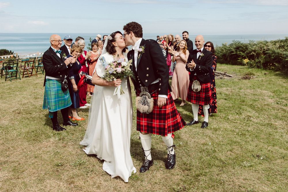 Wedding guests gather outdoors in Scottish kilts and formal attire for group photos on a grassy hillside.