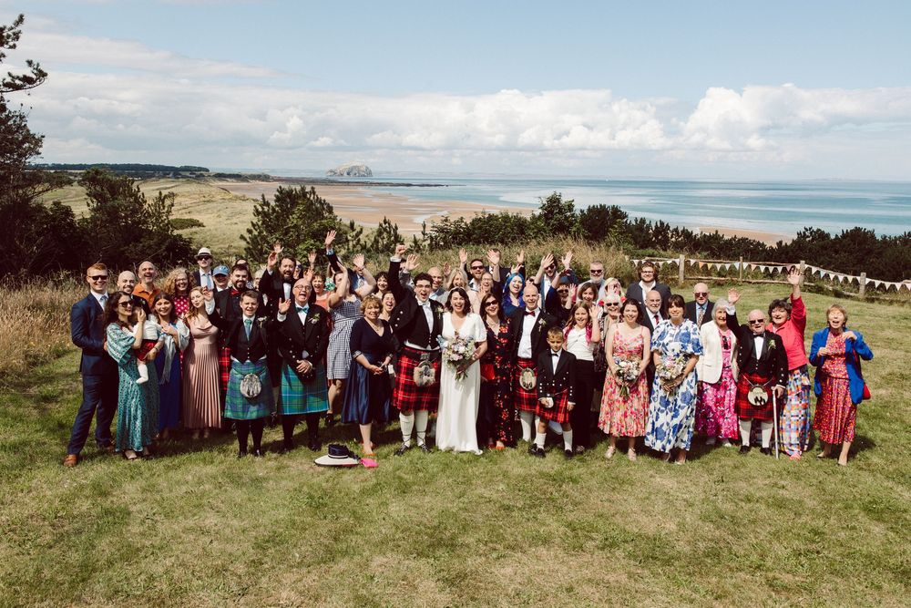Large group of wedding guests pose together outdoors with ocean and blue sky visible in the background.