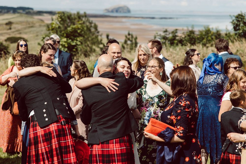 Wedding guests dance and celebrate outdoors in a mix of formal attire and traditional Scottish kilts on a sunny day.