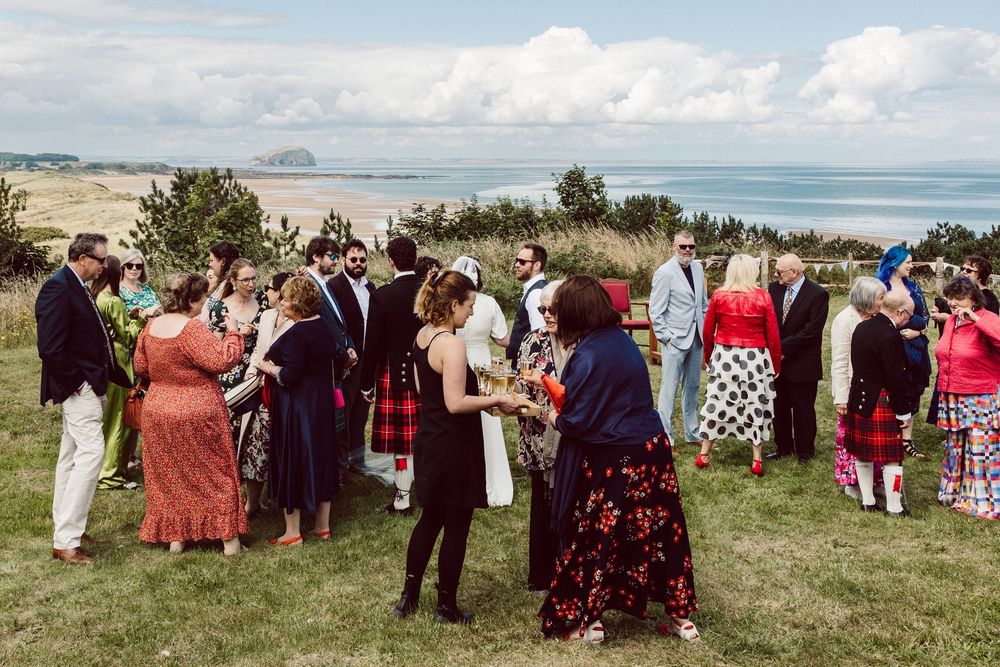 A panoramic view of guests gathered at an outdoor wedding ceremony with the ocean and coastline in the background.