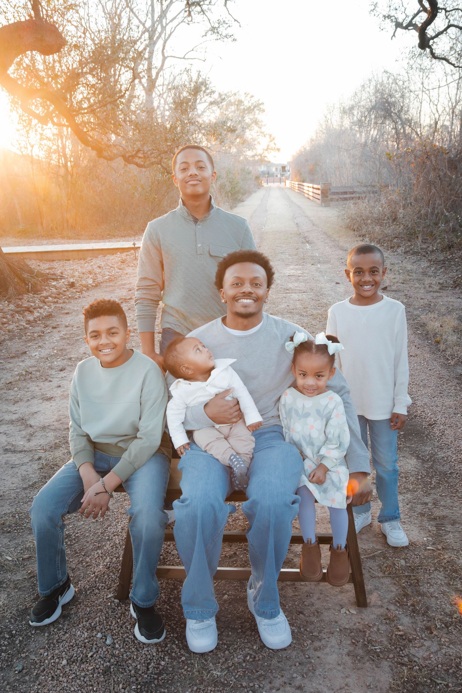 Family sitting together on wooden bench in nature during golden hour sunset wearing coordinated blue and gray outfits.