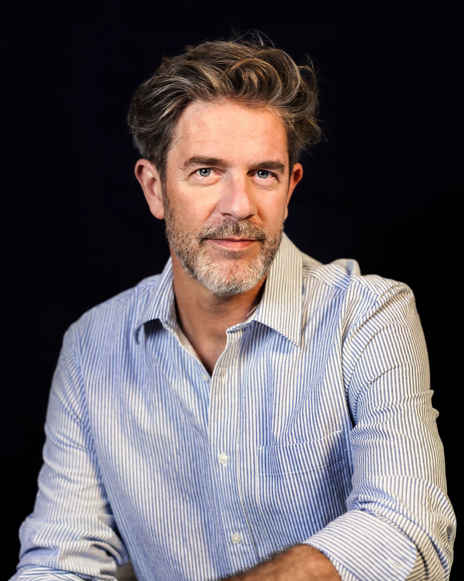 Professional studio portrait of someone wearing a blue and white striped button-down shirt against a dark background.