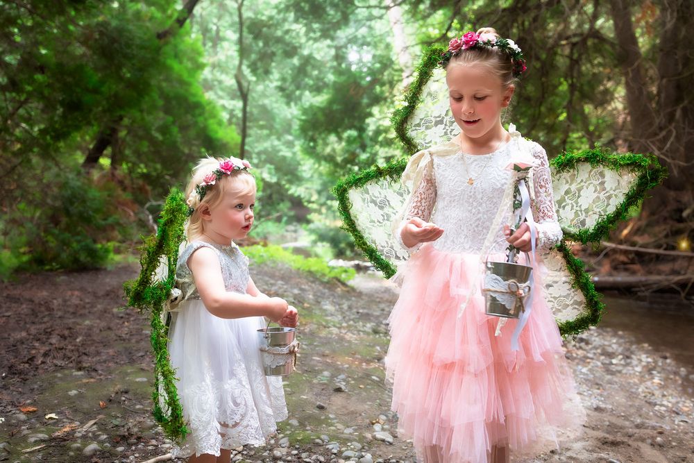 Two children in fairy costumes with wings and floral crowns play together in a magical forest setting.