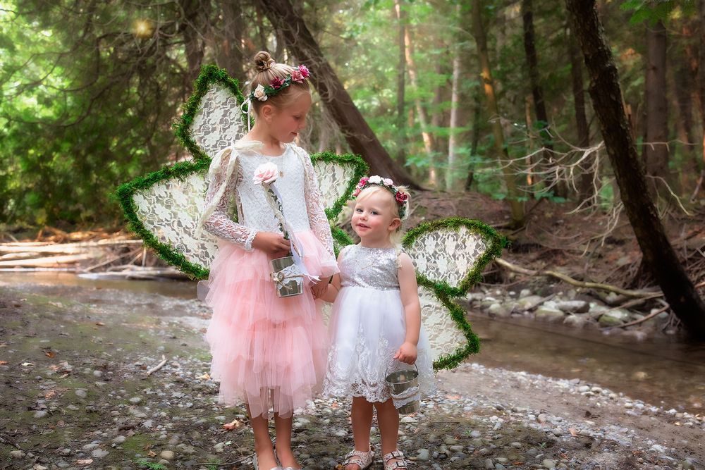 Two figures in pink and white dresses walk hand in hand along a wooded creek bed in a forest setting.