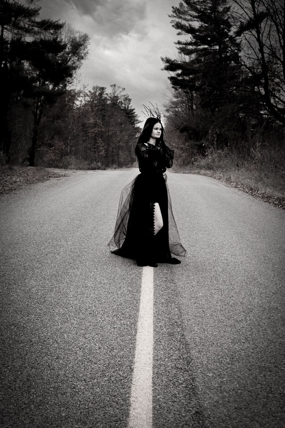 A dark silhouette stands on an empty road with white dividing line under moody cloudy skies in black and white.