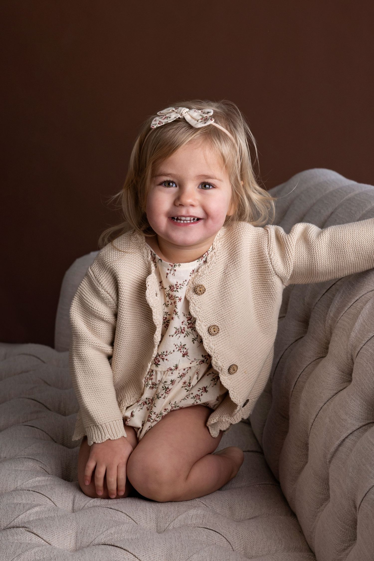 A series of four photos showing a small child in a beige cardigan and floral dress posing on a brown couch.