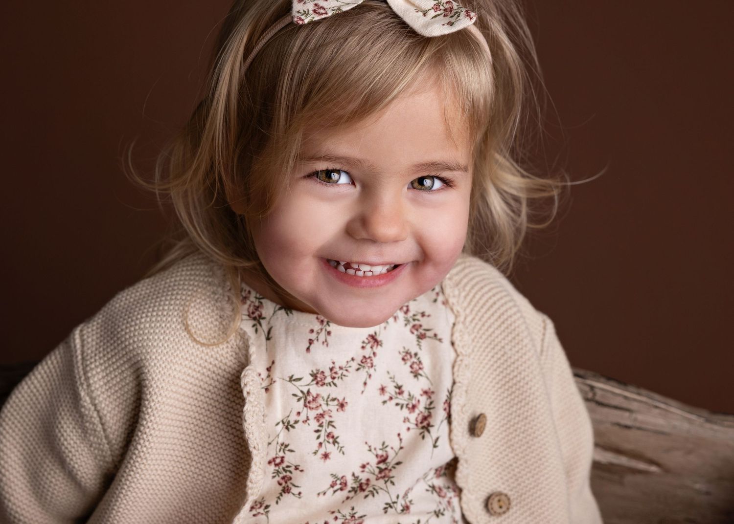 A young child wearing a beige cardigan and floral dress smiles warmly against a brown background.
